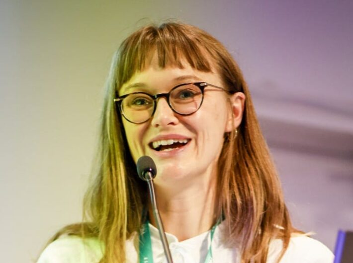 A woman with glasses, long straight hair and a fringe is speaking into a microphone and smiling
