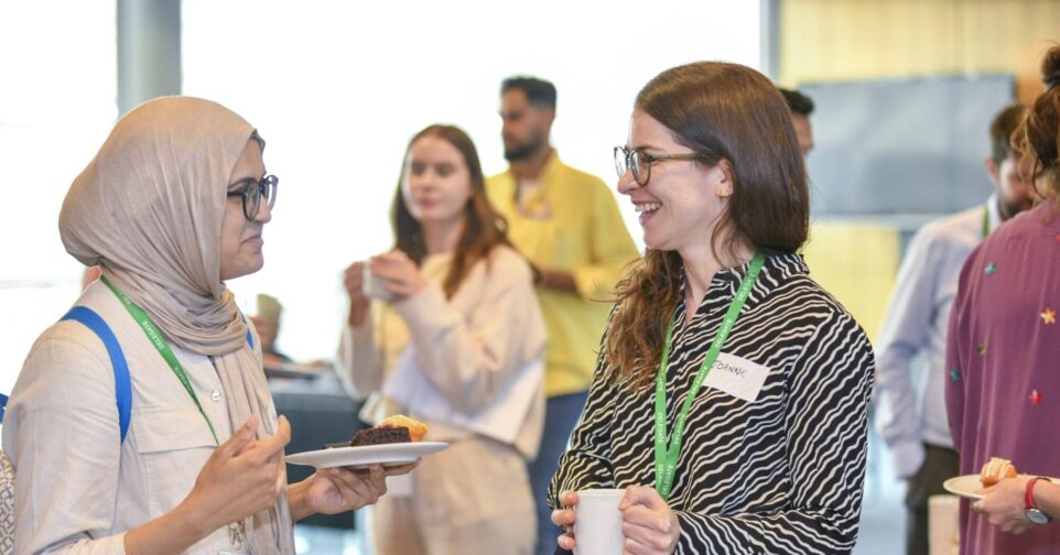 Two people are standing in the foregrand. They are looking at each other and smiling. There are people in the background.