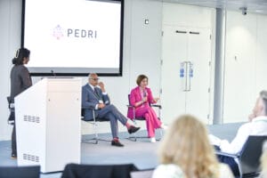 2 people sit down on chairs on a stage. One is in a pink suit holding a microphone and talking. 