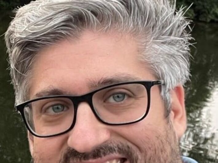 A man with black rimmed glasses, grey stubble and short grey hair smiles at the camera.