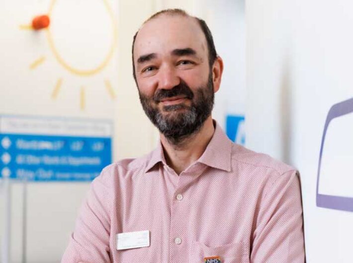 A person with short dark hair and a bear is standing smiling at the camera with their arms crossed. They are wearing a pale red shirt.