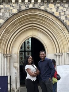 Two people stand in front of a large stone building entrance with a pointed Gothic-style archway. The building façade is made of light stone and flint, and stained-glass windows are visible through the open doorway behind them. A signboard stands to the right of the entrance.