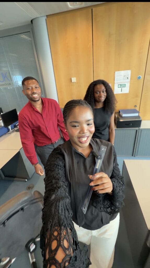 Three people stand in an office space between desks and filing cabinets. One person in the foreground holds a clear award or plaque toward the camera, while two others stand behind. The office contains computer workstations, cabinets, and a glass partition with partial HDR UK branding visible.