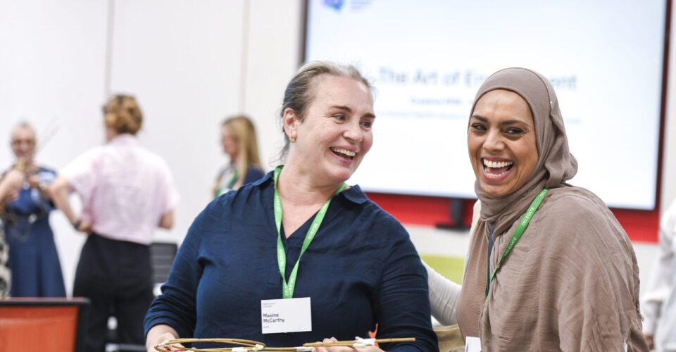 Two conference attendees wearing green event lanyards stand together in a bright meeting room during a networking activity. One person holds a lightweight structure made from thin wooden sticks while both participants take part in a group exercise. Other attendees are visible in the background, and a presentation screen is displayed behind them.