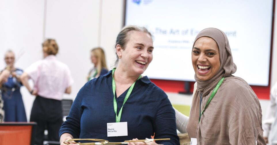 Two conference attendees wearing green event lanyards stand together in a bright meeting room during a networking activity. One person holds a lightweight structure made from thin wooden sticks while both participants take part in a group exercise. Other attendees are visible in the background, and a presentation screen is displayed behind them.